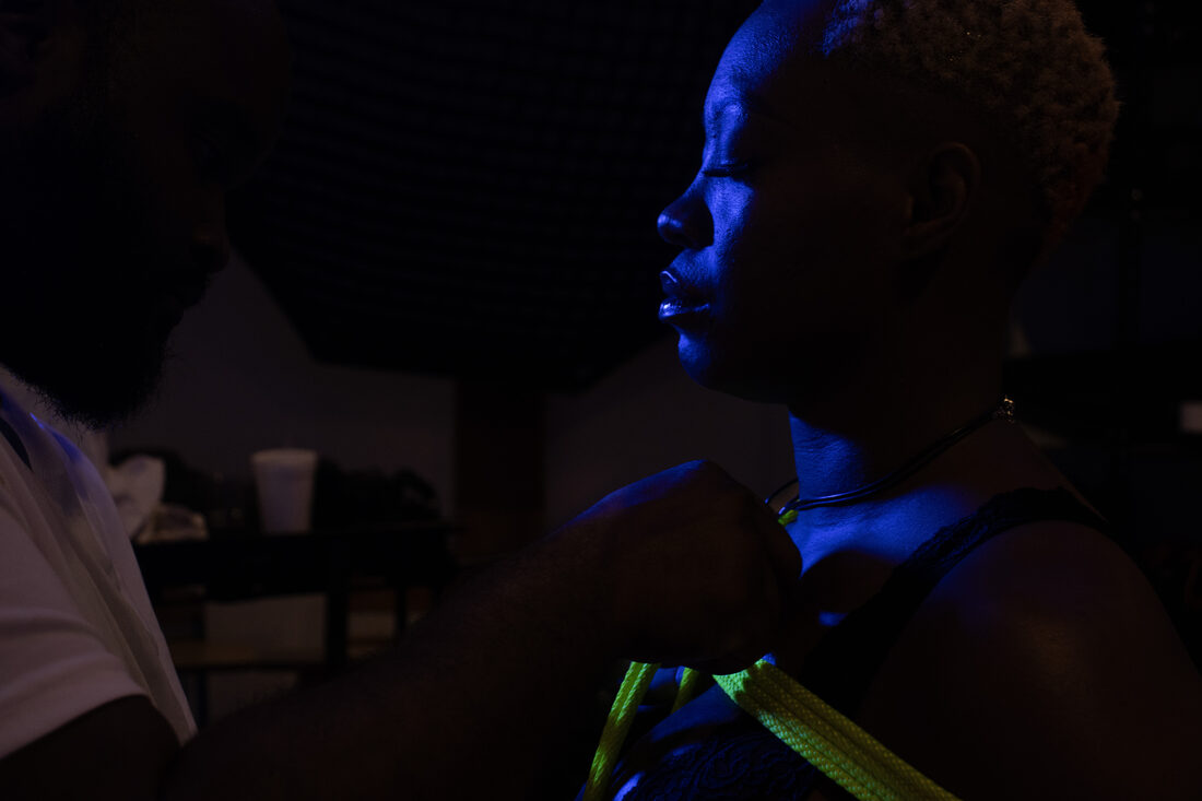 Profile portrait under deep blue light, neon yellow rope at the chest.