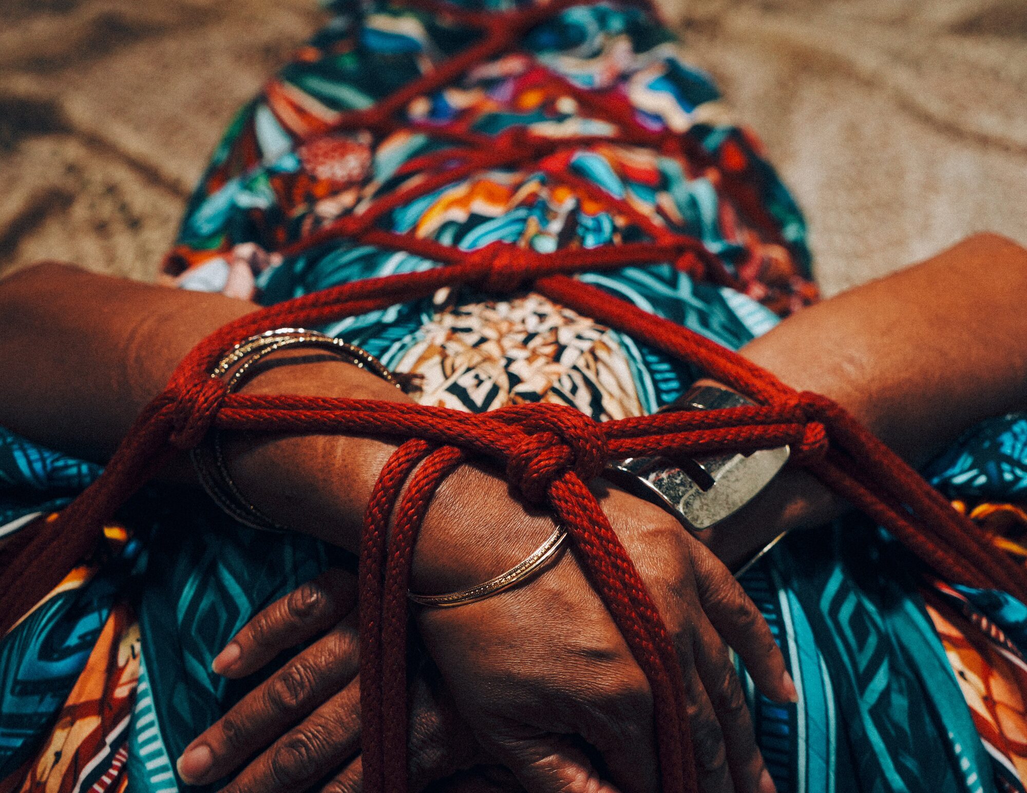 Hands bound together in red rope resting over the chest, body in repose on patterned cloth, the quiet after the container has been built