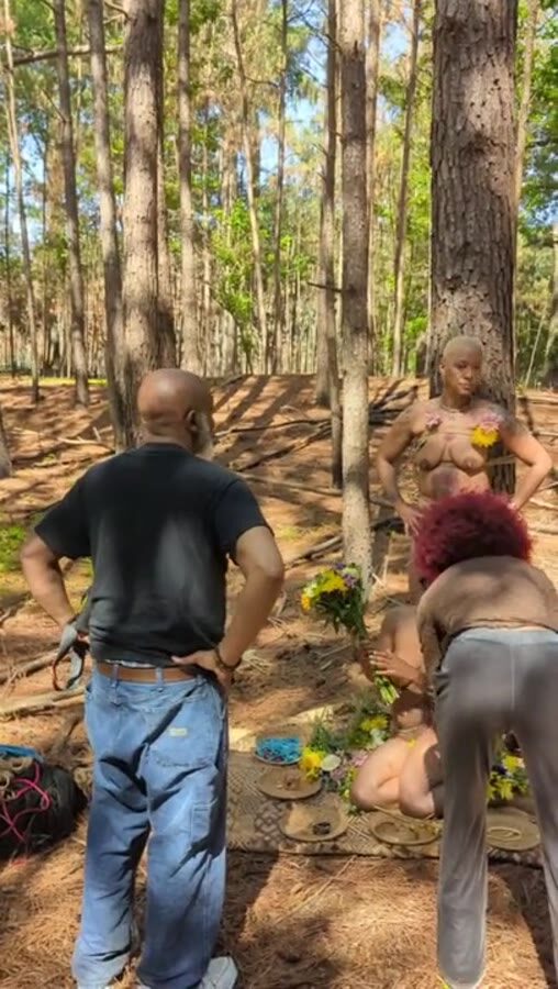 A standing figure in the forest while a collaborator arranges blooms at her feet.