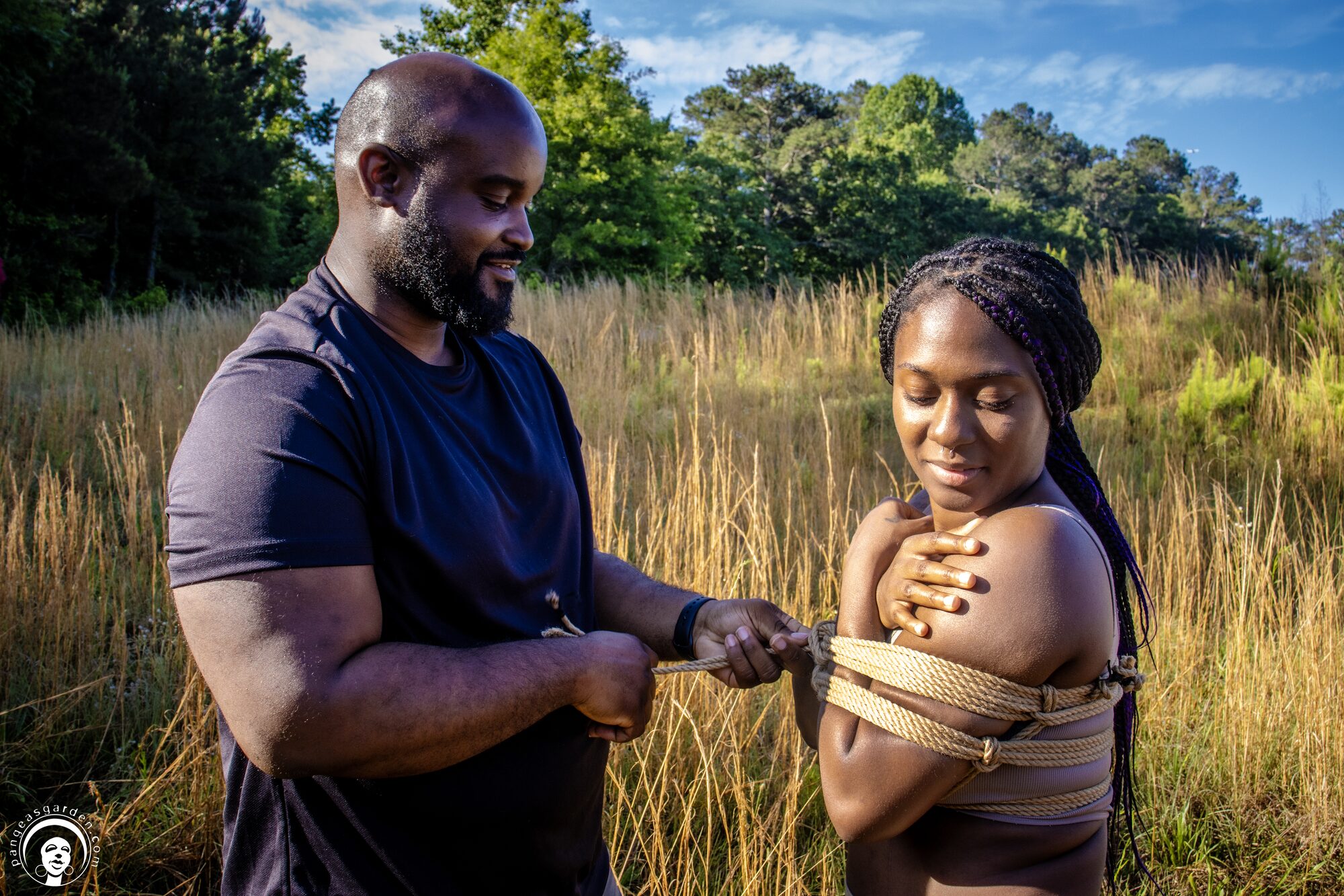 A rigger ties a chest band on a focused recipient who stands waist-deep in tall grass.