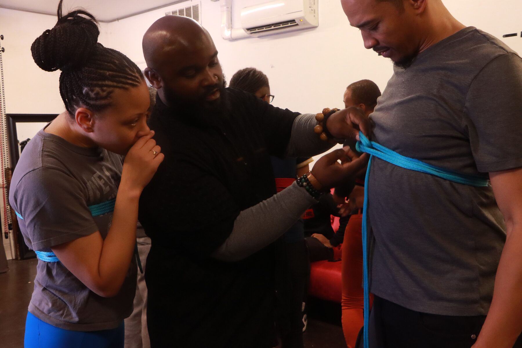 Craig tying a teal chest harness on a standing student while a second student observes at his shoulder