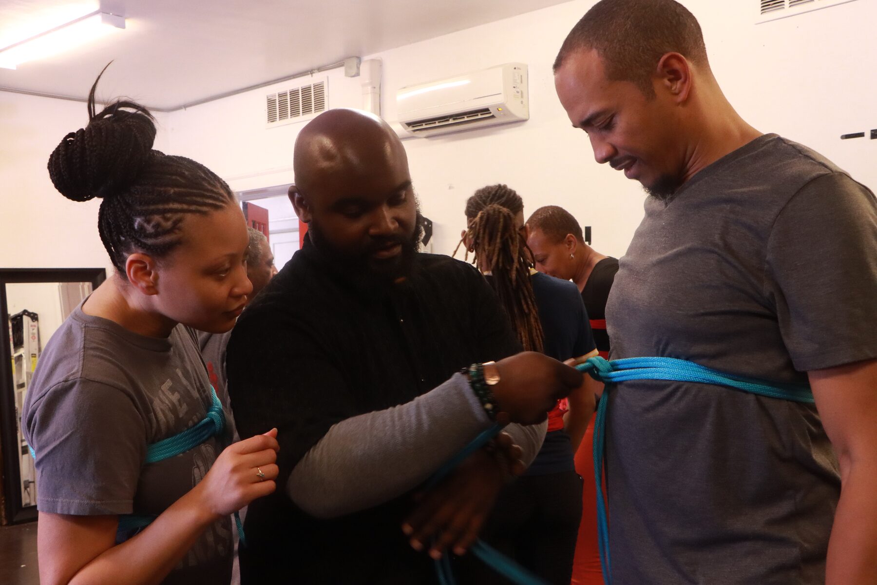 Craig guiding and explaining a teal chest harness tie, hands on the rope, a student watching